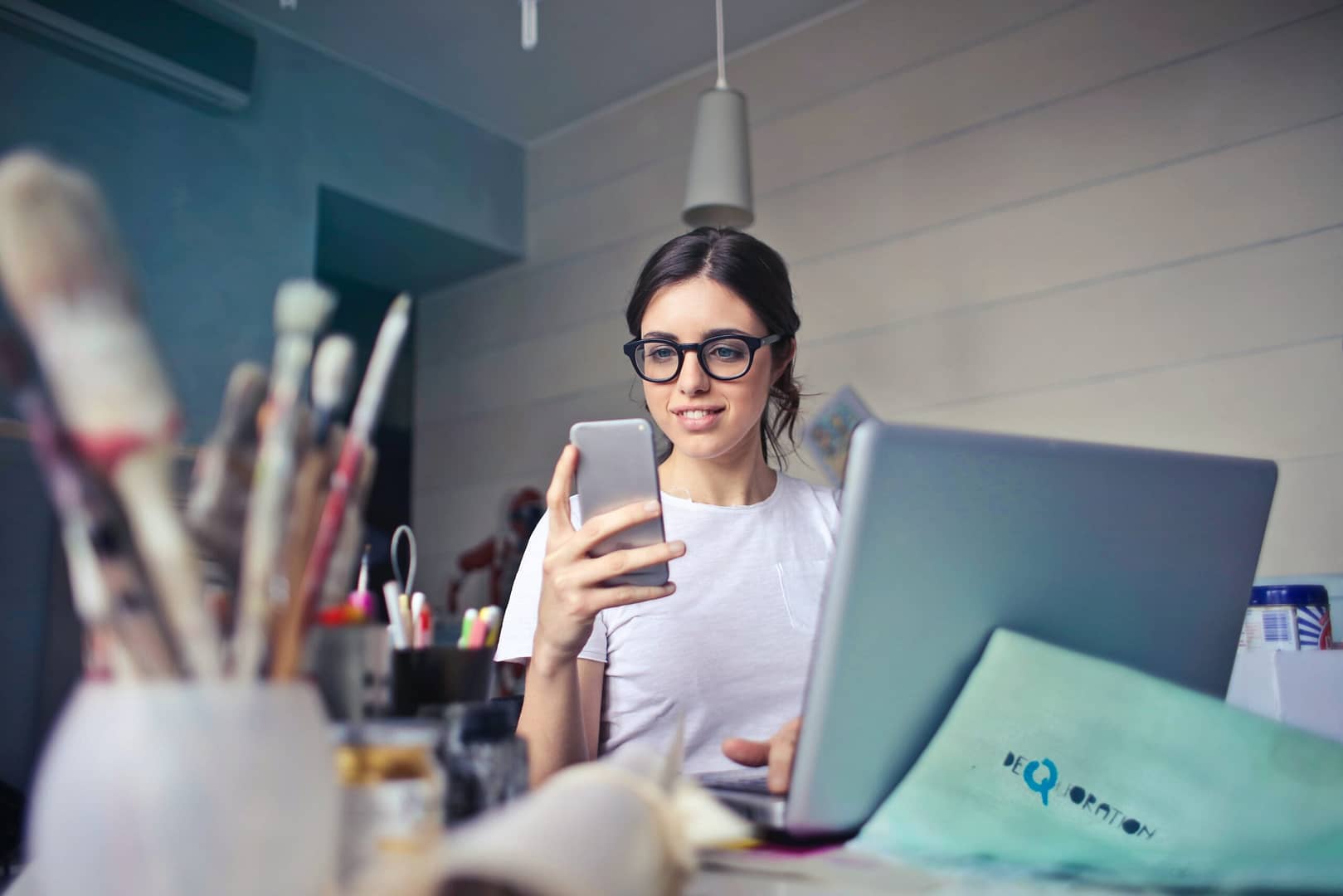 Focused businesswoman multitasking on her laptop and phone, dedicated to overtime work for strategic business planning. Surrounding her, a creative workspace with a laptop, phone, and art brushes, showcasing a blend of technology and artistic inspiration.