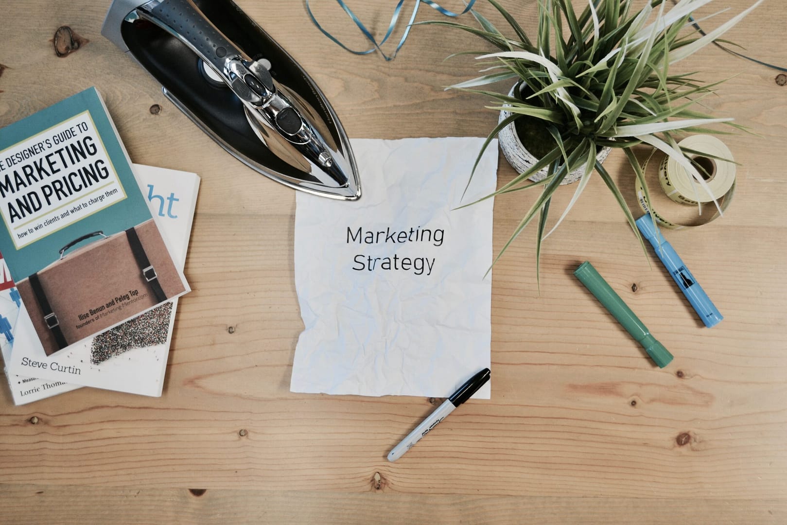 An organized table arrangement featuring a paper titled 'Marketing Strategy' surrounded by potted plants, writing pens, and marketing books