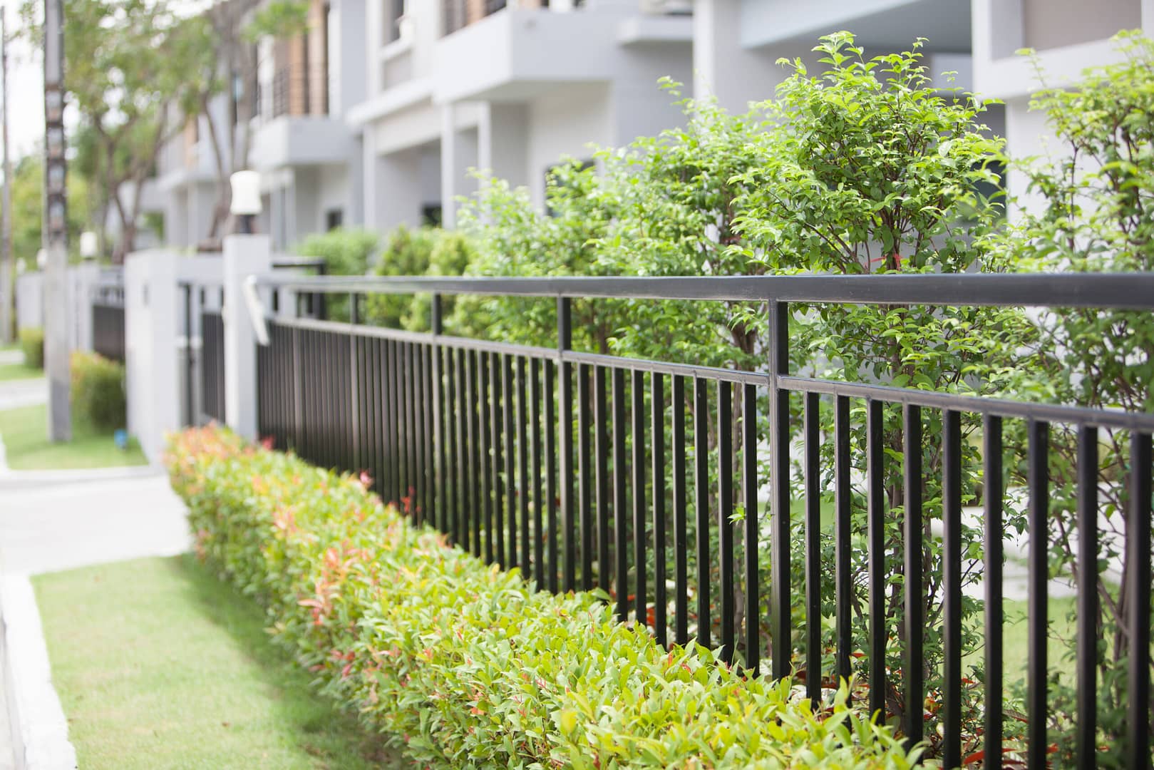 Black fence providing security to a house, framed by lush green trees and neatly trimmed bushes.