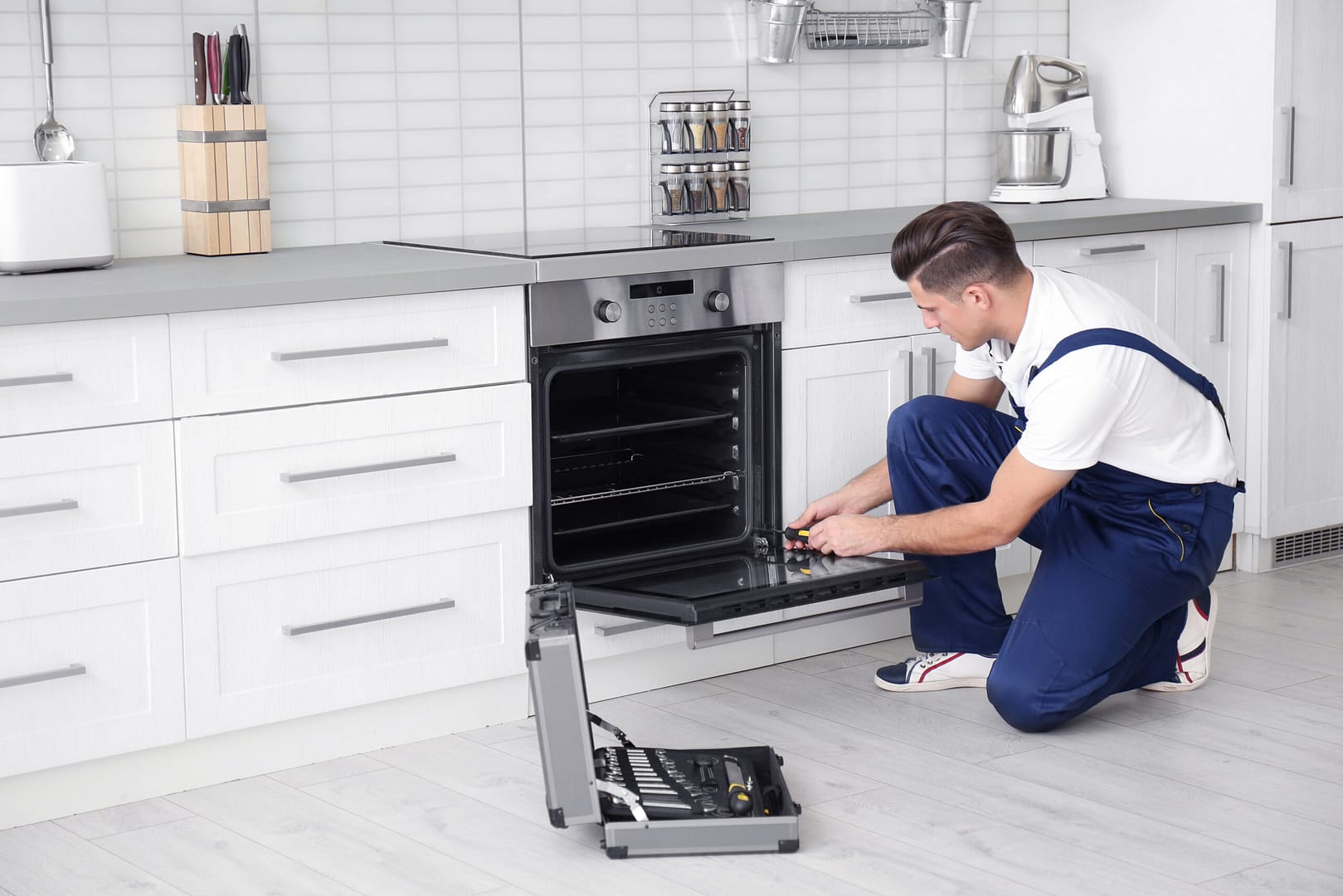 A young man wearing a blue shirt and jeans, focused on repairing an oven in a well-lit kitchen with tools and parts spread out on the counter.