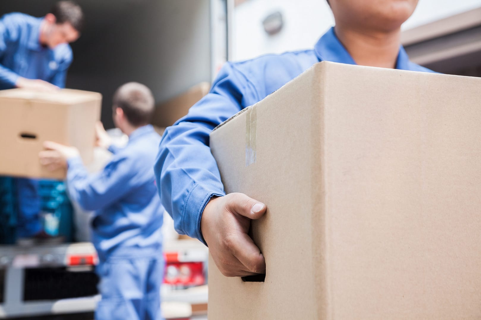 A group of movers in blue uniforms unloading furniture and boxes from a moving van onto the driveway.