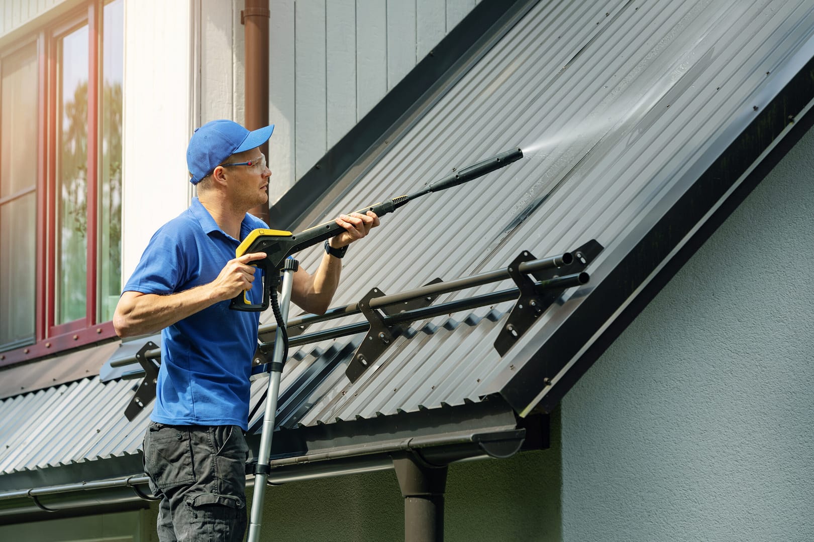 man standing on ladder and cleaning house metal roof with high p