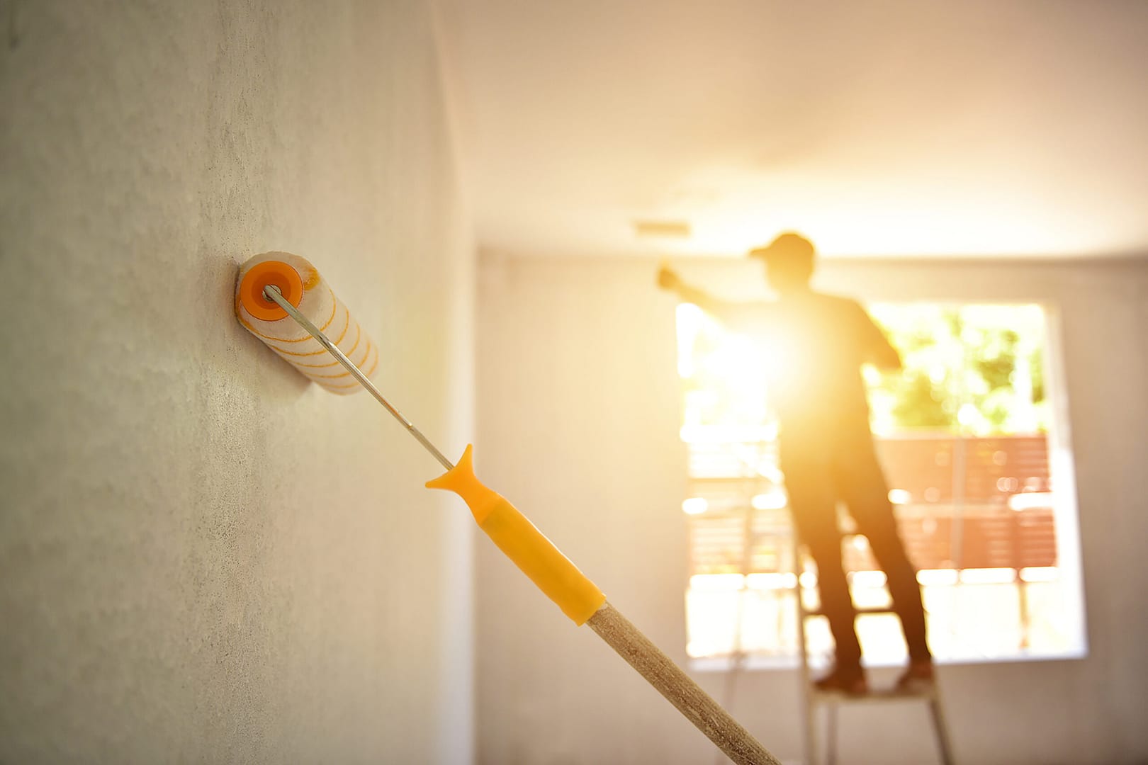 An artist using a paint roller to coat the wall with fresh paint, while a man on a ladder in the background diligently paints the ceiling.