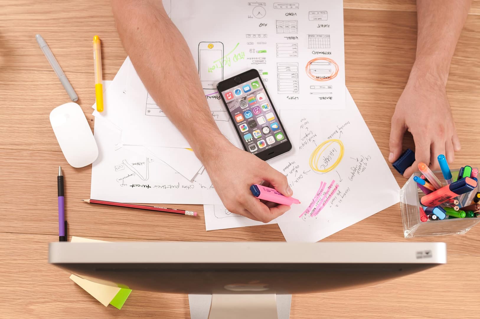 Man diligently writing on paper with a highlighter, smartphone resting atop papers, all set before a computer monitor.