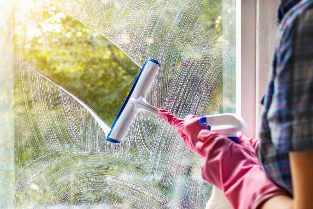 A woman wearing pink gloves diligently cleans the interior of a window using a squeegee, ensuring a spotless, streak-free finish.