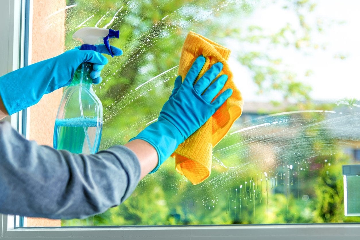 Hands in blue gloves meticulously cleaning the interior of a window using a spray bottle and a vibrant yellow cloth.