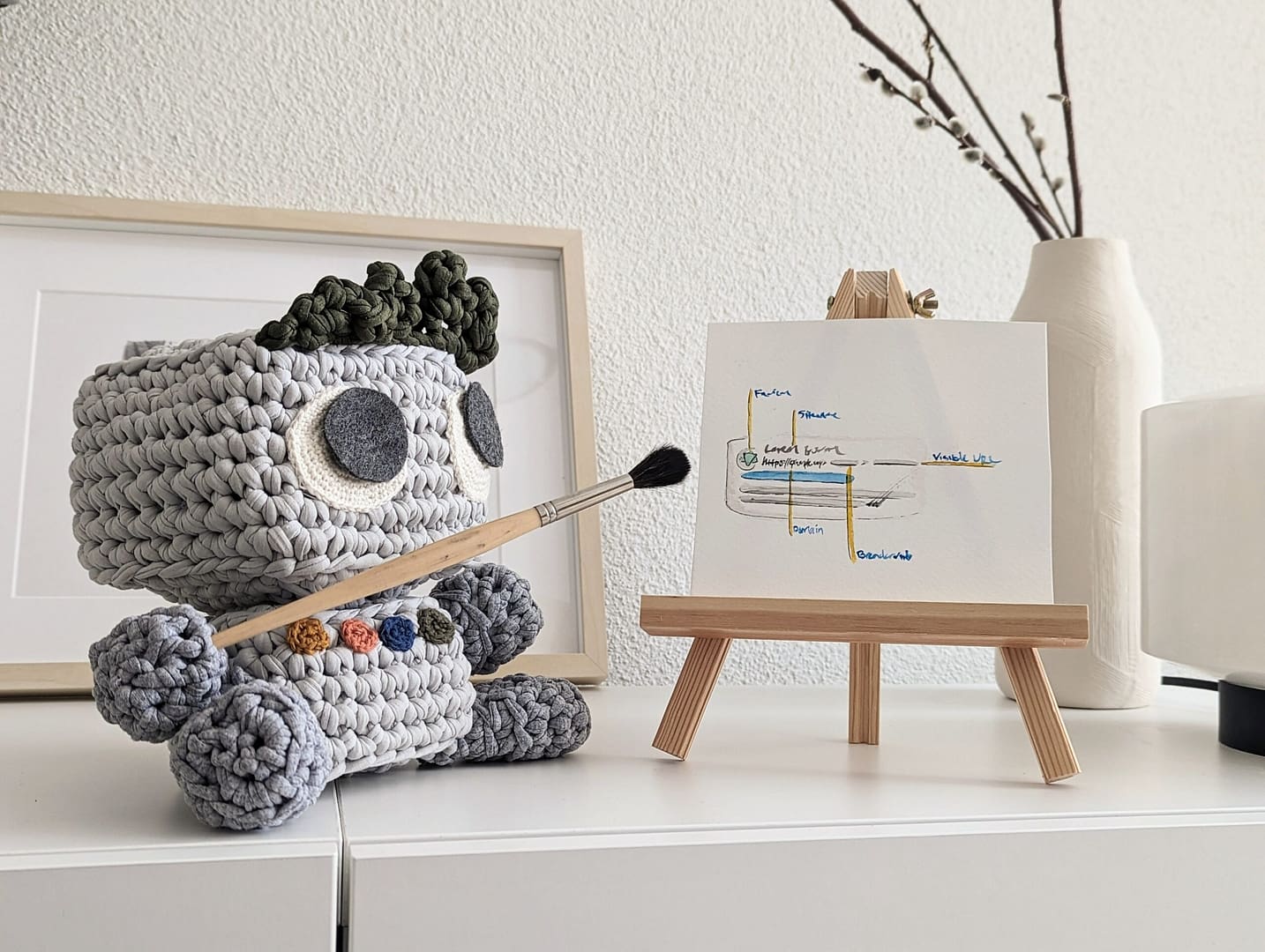 Adorable knitted stuffed toy artist, holding a paintbrush and enthusiastically pointing it at a piece of paper covered in creative plans and designs.