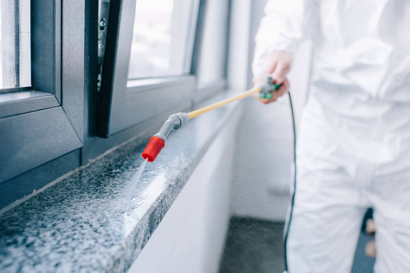 Pest control worker wearing protective gear spraying pesticides on a windowsill at a home to eliminate pests.