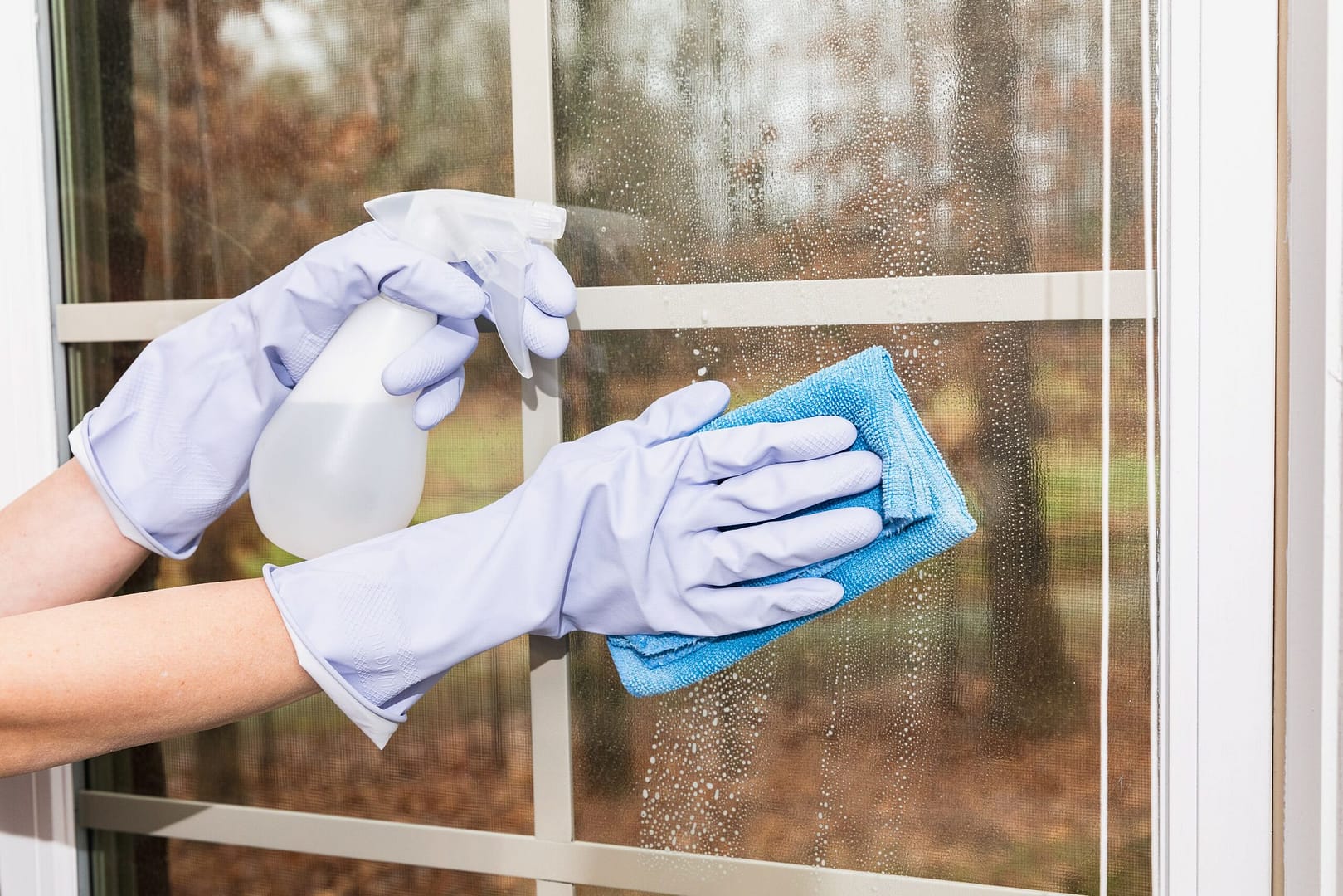 Person using a spray bottle to apply cleaning solution on the outside of a window, followed by wiping it clean with a cloth.