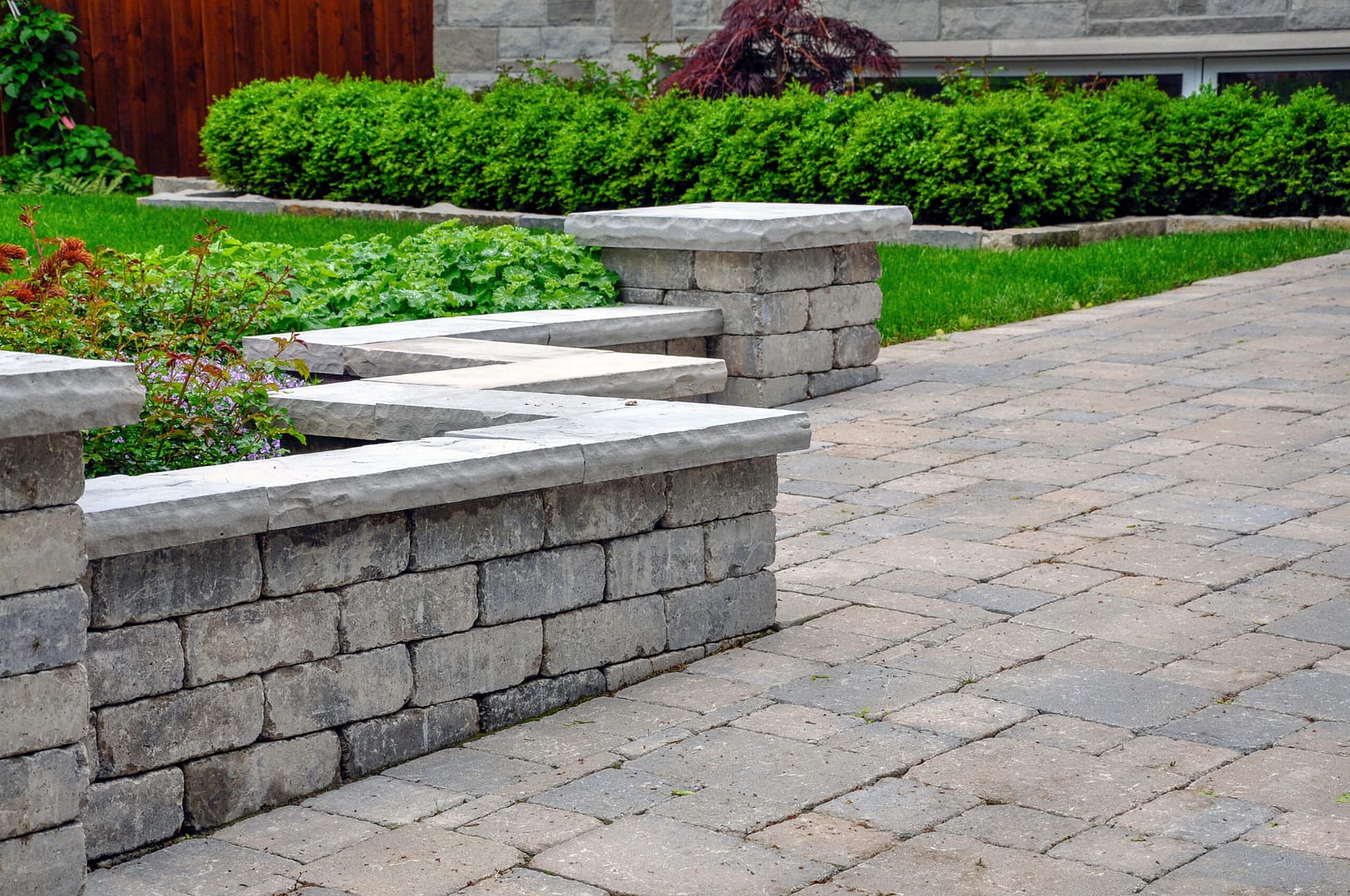 An elegant seat wall featuring sturdy pillars and natural stone coping, defining a charming tumbled paver driveway, creating a picturesque landscaping focal point.
