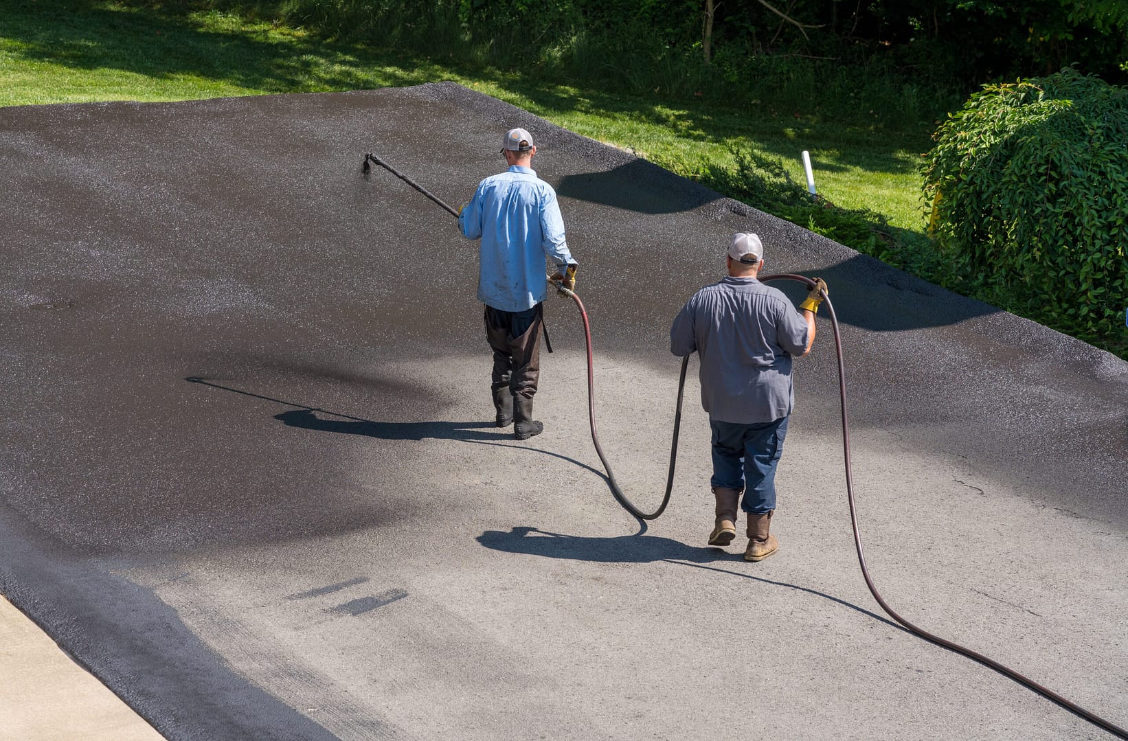 Workers using a spray machine to apply blacktop sealer to an asphalt street, creating a protective coat against the elements.