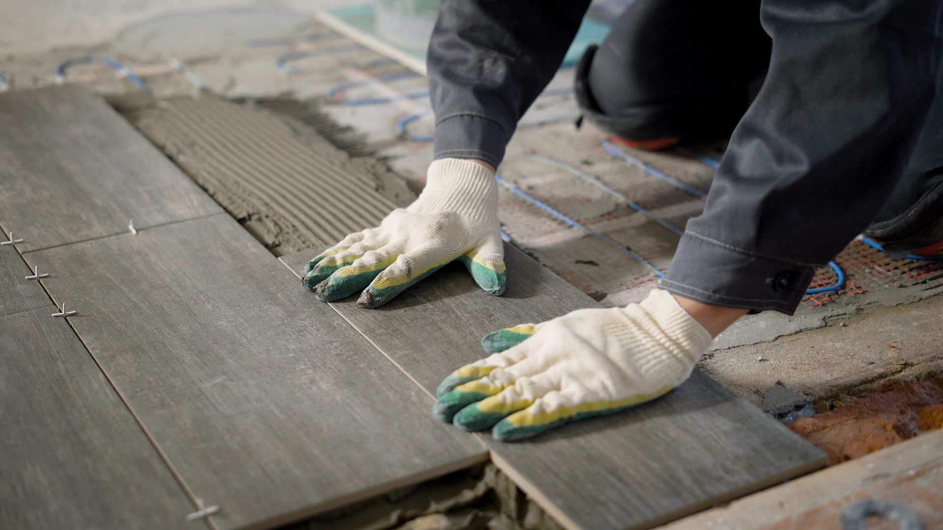 A tiler carefully aligns ceramic floor tiles above a cement surface, with electric floor heating underneath, using a building level to ensure precision in this modern repair project.